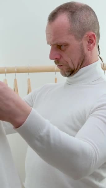 man against background of wooden hanger with white clothes and grimaces trying on putting shirt on himself he lowered corners of lips he doesnot  - Footage, Video