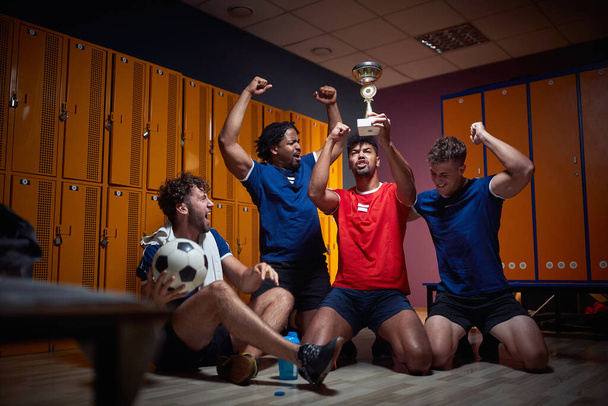Young team players celebrating first place won, holdin golden trophy and cheering in the locker room. Football players feeling victorious. - 写真・画像