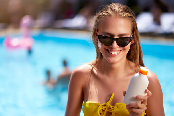 Blonde woman applies sunscreen solar cream on cheeck and face near swimming pool. Smiling pretty girl puts suntan cream from plastic bottle on skin in spa. Female in bikini with a suntan lotion. - Fotó, kép