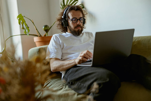 ヘッドフォンの男は自宅で居心地の良いソファに横たわって、ラップトップを使用しています。距離作業概念  - 写真・画像