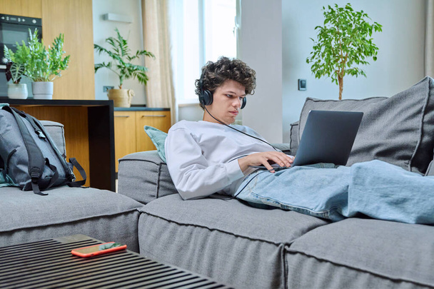 Young guy 18-20 years old resting at home, lying on sofa in living room, looking at laptop screen in headphones. Internet online technology, study leisure communication concept - Foto, Imagen