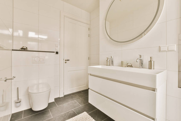 Lavabonun üstündeki duvarda siyah döşemeli beyaz bir banyo ve yuvarlak bir ayna oval şekilli bir ayna. - Fotoğraf, Görsel