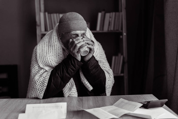 Vanha mies jäätyy kotona istuu pöydän ääressä tarkistamassa lämpö- ja sähkölaskuja, juomassa kuumaa teetä pysyäkseen lämpimänä, ei tarpeeksi rahaa lämmitykseen. Mustavalkoinen - Valokuva, kuva