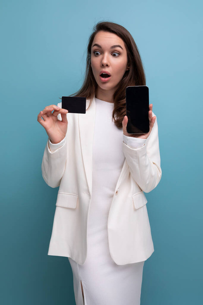 nette brünette junge Frau in Kleid und Jacke mit einer Bezahlplastikkarte mit einem Smartphone. - Foto, Bild
