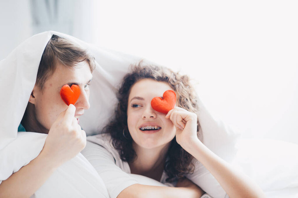 Love couple lying on bed under blanket holding red hearts, happy romantic life - Photo, Image