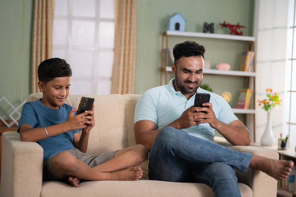 focus on father, Indian father and son busy on there mobile phones while sitting on sofa at home - concept of technology addiction, social media and togetherness. - Фото, изображение