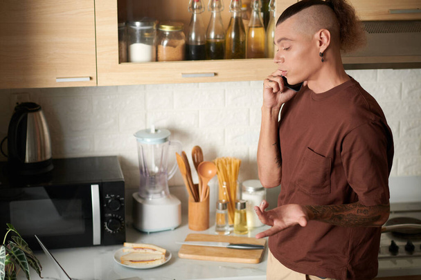 Νεαρός μιλάει στο τηλέφωνο με συνάδελφο όταν μαγειρεύει πρωινό στην κουζίνα - Φωτογραφία, εικόνα