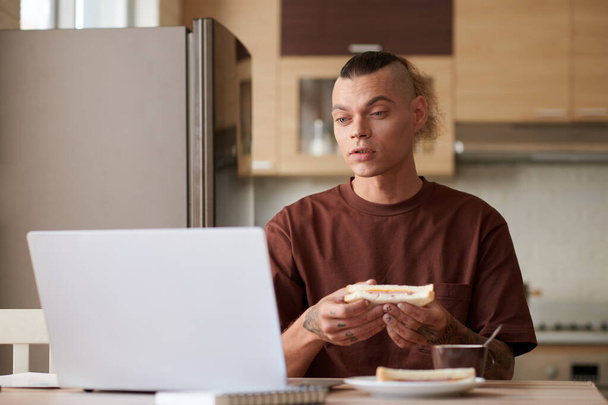 Młodzieniec jedzący kanapkę z szynką i serem podczas oglądania smyczy na laptopie - Zdjęcie, obraz