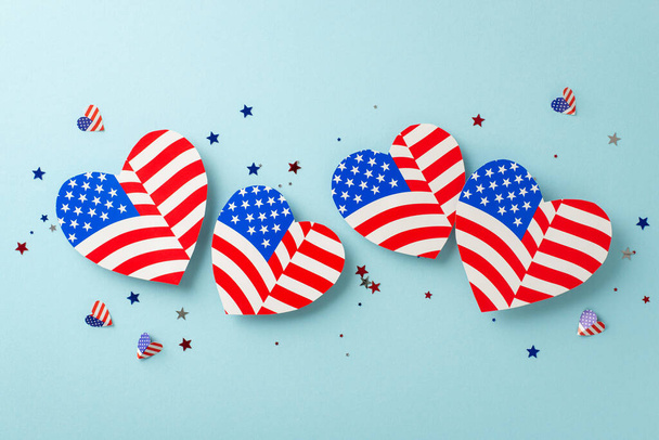 Festas do Dia da Independência dos EUA. Da vista superior, olhe para adornos emblemáticos: corações com o motivo da bandeira americana, confetes brilhantes em um design temático, exibidos em pano de fundo azul pastel - Foto, Imagem