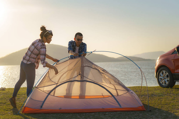 Asyalı çift gün batımında arka planda gölle birlikte çimenlikte kamp yapmak için çadır kuruyor. - Fotoğraf, Görsel