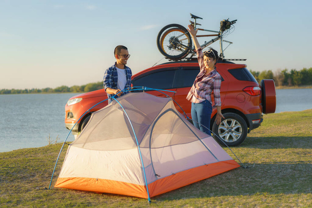 Couple asiatique préparant une tente pour camper dans la pelouse avec le lac en arrière-plan pendant le coucher du soleil - Photo, image