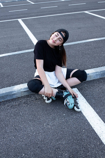 Cheerful girl in roller skates sitting in an open parking lot posing. Stylish clothes. Caucasian teenager, in a good mood. Outside. Urbanism, hobby, roller skating, holidays, city concept. Day light. - Фото, изображение