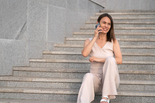 Μια νεαρή γυναίκα κάθεται στα σκαλιά μιας σκάλας έξω και μιλάει σε ένα κινητό τηλέφωνο. Υψηλής ποιότητας φωτογραφία - Φωτογραφία, εικόνα