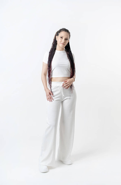 Modieuze jonge mooie vrouw. Slank meisje met dreadlocks in een actieve pose in een witte broek en top. Mode, kleding en stijl. - Foto, afbeelding