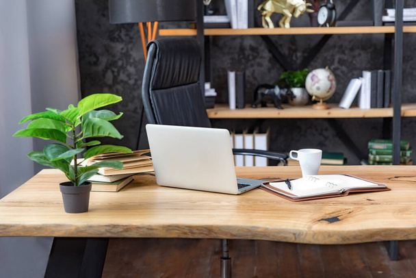 Fragment of the office. Desk with notebook, notepad, pen and plant. Nearby is a beautiful black leather armchair. In the background are shelves with books, globes, clocks and other decor. - Photo, Image