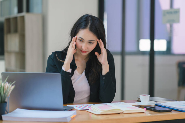 La donna d'affari asiatica millenaria stressata ed esausta è vista seduta alla scrivania del suo ufficio con la mano sulla testa, indicando una dura giornata di lavoro in cui è sovraccarica di lavoro. - Foto, immagini