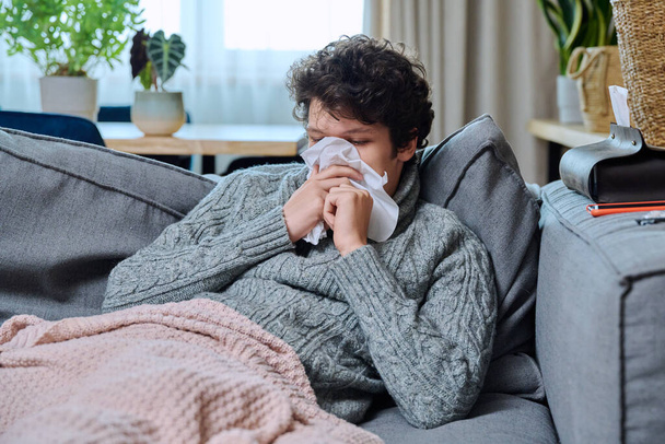 Sick young guy sneezing into nasal tissue, having signs of respiratory infectious disease, in woolen sweater sitting on couch at home. Runny nose, cough, cold season - Photo, image