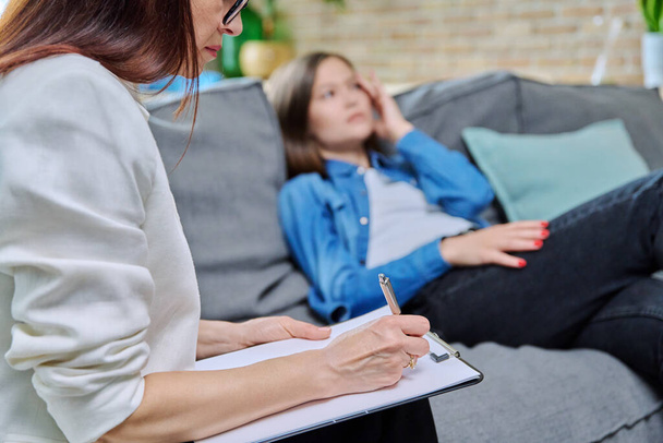 Close-up of female psychologists hands with clipboard making notes, in individual therapy session. Young women patient on couch out of focus. Psychology psychotherapy treatment mental health concept - Foto, immagini