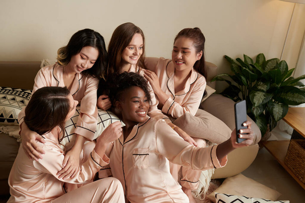 Улыбающаяся счастливая молодая женщина в шелковой пижаме делает селфи с друзьями - Фото, изображение
