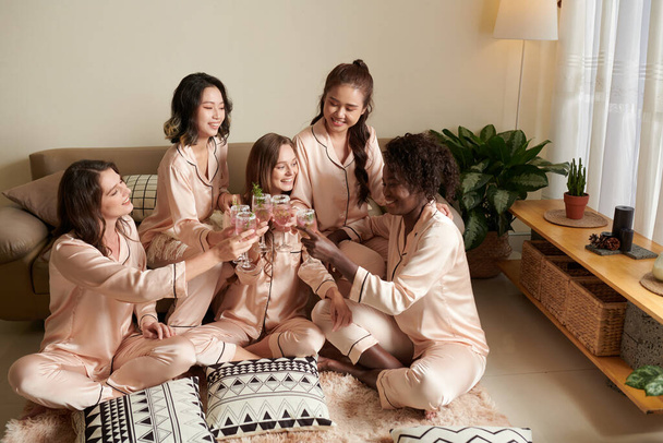 İpek pijamalı bir grup arkadaş yatak odasında yerde oturmuş margarita bardaklarıyla kadeh kaldırıyorlardı. - Fotoğraf, Görsel