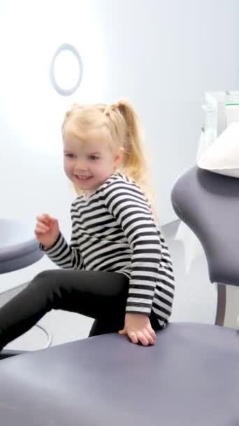 маленькая девочка в стоматологическом кабинете самостоятельно забирается на место для стоматологического лечения стоматологическое кресло с игрушкой по новейшим технологиям для лечения детского молока  - Кадры, видео