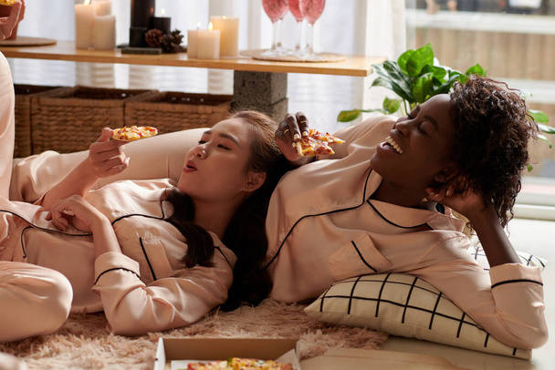 Mujeres jóvenes felices en pijama descansando en el suelo, apoyándose unas sobre otras y comiendo rebanadas de deliciosa pizza - Foto, Imagen