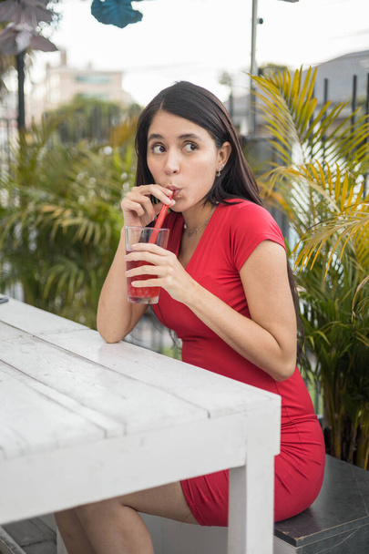 vonzó fiatal latin nő élvezi az italt a kertben nappal, visel piros ruhát, szépség és alkalmi divat, természetes és egészséges gyümölcslevek - Fotó, kép