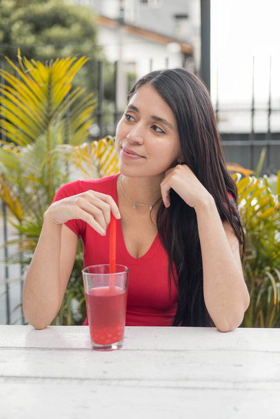 cheerful beautiful woman enjoying a drink in a garden on a summer day, she wears a red dress, beauty and casual fashion, natural and healthy juices, lifestyle - Photo, Image