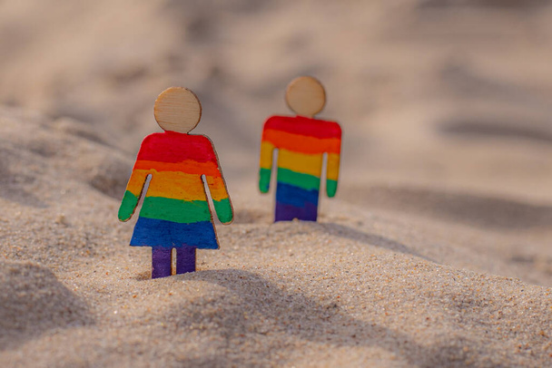 Persone LGBT in legno sulla spiaggia sulla sabbia. Coppia in piedi sulla spiaggia con bandiera arcobaleno, pari diritti, simbolo della comunità LGBT. Bandiera LGBT su una spiaggia sabbiosa. Concetto di viaggio estivo. Simbolo dello stesso sesso - Foto, immagini