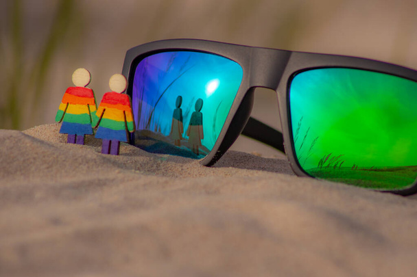 LGBT dřevěné lidi na pláži na písku. Pár stojící na pláži s duhovou vlajkou, rovná práva, symbol komunity LGBT. Sluneční brýle s vlajkou LGBT na písečné pláži. Koncept letního cestování. - Fotografie, Obrázek