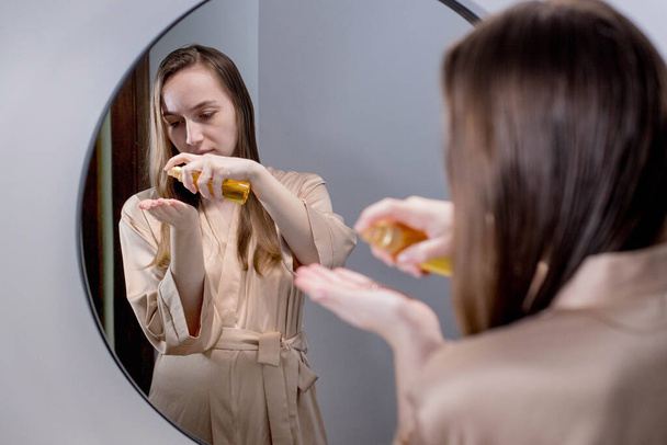 Yong mulher morena aplicando spray de óleo essencial em seu cabelo de pé perto do espelho no banheiro. Dividir termina o tratamento de reparação. Conceito de proteção e cuidado para o cabelo. - Foto, Imagem