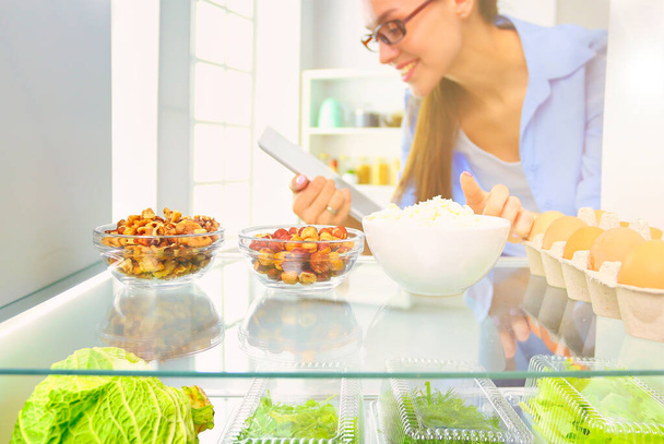 Portrait of female standing near open fridge full of healthy food, vegetables and fruits - Foto, Bild