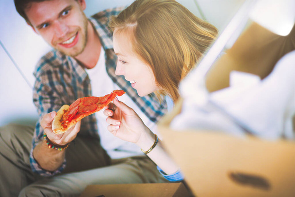 Ένα νεαρό ζευγάρι κάνει ένα διάλειμμα για πίτσα στο πάτωμα αφού μετακομίσει σε ένα νέο σπίτι με κουτιά γύρω του. Νεαρό ζευγάρι. - Φωτογραφία, εικόνα