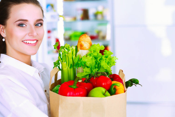 Young woman holding grocery shopping bag with vegetables .Standing in the kitchen. Woman in the kitchen looking at the camera. - Foto, imagen