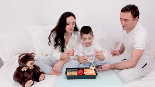 familia comiendo sushi en la cama en casa pidiendo comida a domicilio marido esposa y niño juguete osito de peluche sentado en sábanas blancas niño estudiando comer con palillos delicioso sushi apetitoso feliz vida familiar - Metraje, vídeo