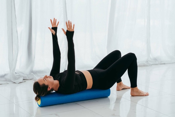 Mujer entrenadora de fitness en ropa negra de fitness en la sala de entrenamiento flexiona con un rollo de masaje en el suelo en la clase de pilates.  - Foto, imagen