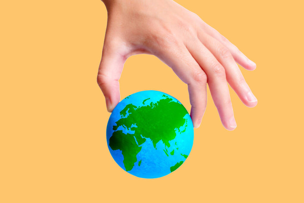 Close-up view of a hand pinch-holding a green and blue model of the planet Earth isolated on yellow background. Creative environmental awareness related concept. - Foto, Imagem