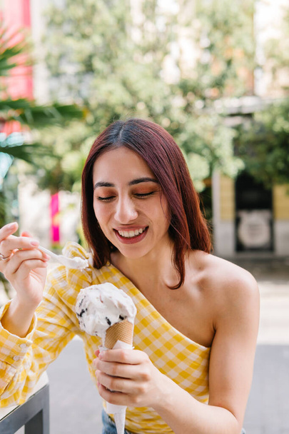Pionowe zdjęcie stylowej kobiety jedzącej stożek lodów na świeżym powietrzu - Zdjęcie, obraz