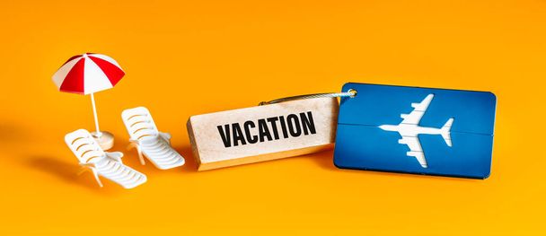Vacation concept, Plane luggage tag, Sun loungers and umbrella miniature, Beautiful yellow background, Panorama, Written vacation on a wooden block - Photo, Image