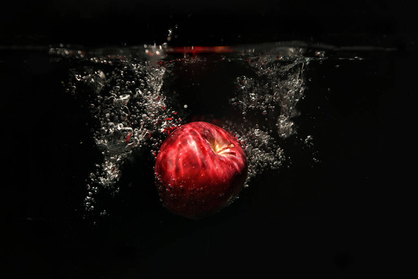 Słodkie czerwone jabłka wpadają do wody z pluskiem i bąbelkami powietrza na czarnym tle. - Zdjęcie, obraz