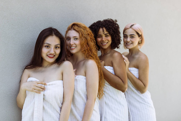 Four girls of different races, dressed in beige towels, stand against a grey wall. Beauty of diversity, SPA experience and multiethnic female friendship concept. Focus on Chinese (first) girl. - Foto, Bild