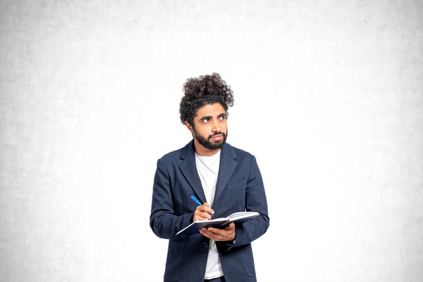 Pensive young Middle Eastern man college student standing with notebook and pen and looking sideways and upwards over concrete background. Concept of planning, creativity and education - Zdjęcie, obraz