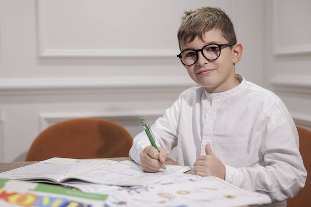 Blond boy in glasses and a blue polo shirt does his homework in his room - Φωτογραφία, εικόνα