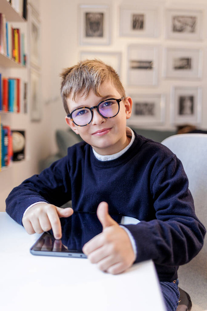 Gözlüklü sarışın çocuk, mavi giyinmiş ve masanın önünde oturuyor. Çalışma odasında arka planda kitaplık olan bir tablet kullanıyor.. - Fotoğraf, Görsel