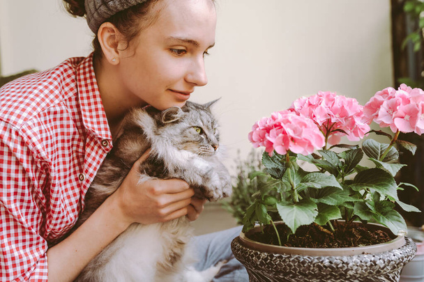 όμορφη ευτυχισμένη έφηβη κοπέλα, μαζί με την αγαπημένη γάτα, θαυμάζει το λουλούδι ορτανσία με ροζ πέταλα φυτεμένα σε γλάστρα. Ασχοληθείτε με το αγαπημένο σας χόμπι κηπουρική με το κατοικίδιο ζώο σας - Φωτογραφία, εικόνα