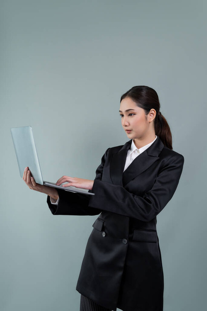 Confident young businesswoman stands on isolated background, working on laptop posing in formal black suit. Office lady or manager with smart and professional appearance. Enthusiastic - Фото, изображение