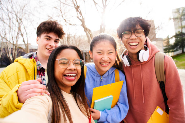 Group of multi-ethnic high school students taking a selfie outdoors at the university campus holding folders. Looking at camera picture of a diverse colleagues having fun.  - 写真・画像