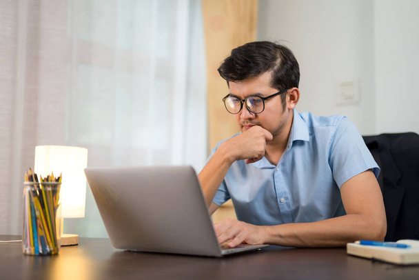 Un joven guapo en gafas usando un portátil en el escritorio leyendo información de redes sociales para mejorar en casa. - Foto, Imagen