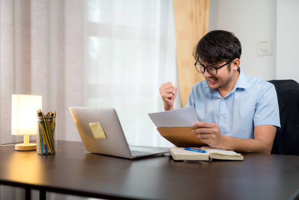 Egy jóképű, szemüveges fiatalember, aki sikeres gesztust mutat, miközben laptoppal az íróasztalán olvassa az elfogadólevelet. - Fotó, kép
