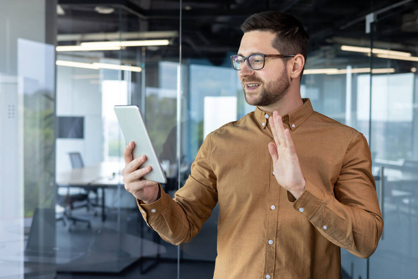 A young businessman man is standing in the office center, holding a tablet in his hands and talking on a video call, greeting and waving at the screen. - Foto, Bild